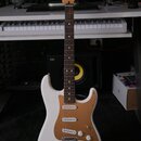 Bílá elektrická kytara typu Stratocaster s hnědým pickguardem a hnedým hmatníkem. Na hlavě je vidět nápis Fender. Kytara má tři snímače, tři ovládací…
