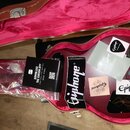 Otevřený hnědý tvrdý kufýr s červeným plyšovým vnitřkem. Uvnitř jsou various doplňky, včetně černého manuálu s nápisem DOWNLOAD THE GIBSON APP a něko…