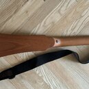 Dřevěný strunný nástroj s tělem ve tvaru pušky v hnědé barvě. Nástroj má úzký krk a hlavu s osmi kovovými ladičkami. K nástroji je přiložen černý tex…