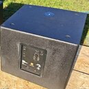 Černý subwoofer s texturovaným povrchem v kostkovém tvaru. Na zadní straně je ovládací panel s několika přepínači a konektory, u kterých jsou vidět n…