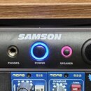 Šedý rackový zesilovač značky SAMSON, model SERVO 120a. Přední panel obsahuje dva ovratidla pro kanály CHANNEL 1 a CHANNEL 2, LED indikátory s nápisy…
