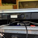 Černý rackový přístroj s nápisem SHURE GLX-D+ umístěný v hliníkovém transportním kufru. Zařízení má digitální displej s textem auto 1A, zapínací tlač…