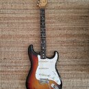 Elektrická kytara typu Stratocaster s barevným přechodem korpusu od černého přes červený k žlutému. Na hlavě hmatníku je viditelné logo Fender. Kytar…