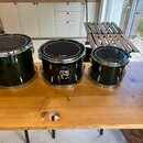 Tři černé bubny s kovovými obručemi a ladidly umístěné na dřevěném stole. Na každém z bubnů je v horní části bílý nápis "Gretsch". Povrch nástrojů je…