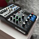 Černý kompaktní mixážní pult značky YAMAHA. Na horní části jsou vidět vstupy a výstupy, nápisy 1/L, 2/R, 3/4 - 5/6 a označení modelu MG06. Přístroj o…