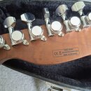 Detail zadní strany hlavy kytary z hnědého dřeva s šesti chromovanými ladicími mechanismy. Na dřevě je černý nápis SERIAL NUMBER, značka CE, piktogra…