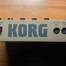 Záběr na zadní panel šedého elektronického zařízení s velkým nápisem KORG. Jsou zde vidět konektory DC 9V, STANDBY ON, MIDI THRU, MIDI OUT a MIDI IN.…