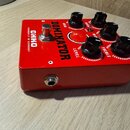 Červený efekt pedál s nápisy DOMINATOR a OHKO HANDBUILT IN GERMANY. Na horní straně se nachází pět černých oválných knoflíků s popisky GAIN, BASS, MI…