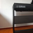Detailní pohled na levou část digitálního pianina v černé barvě. Na horní části je bílý nápis YAMAHA doprovázený logem značky. Nástroj stojí na černé…