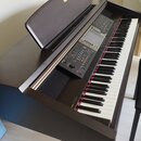 Digitální piano v tmavě hnědém dřevěném korpusu s černou ovládací panelí a bílo-černými klávesami. Na spodní liště je zlatý nápis Yamaha Clavinova a…