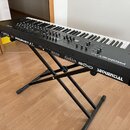 Černý analogový syntetizátor s klaviaturou umístěný na černém X-stojaně. Na předním panelu jsou nápisy PROPHET XL, SEQUENTIAL a obráceně napsaný text…