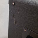 Detailní pohled na roh černého reproduktorového boxu s texturovaným povrchem. V horním rohu je připevněn černý plastový ochranný roh. Povrch v místě…