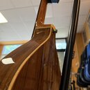 Detailní pohled na zadní část akustické kytary s hnědým dřevěným povrchem a viditnou sletovanou strukturou. Nástroj je umístěn v černém kovovém stoja…