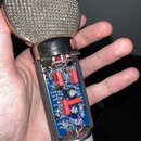 Fotografie znázorňuje mikrofon s odsunutým krytem, který odhaluje vnitřní elektroniku na modré desce s kondenzátory a zapájkami. Na spodní části kovo…