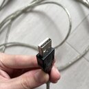 Fotografie zachycuje stříbrný kabel s konektorem USB typu B, který drží ruka. Na černém plastovém krytu konektoru je vyryto symbol USB. Ve spodní čás…