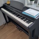 Černý digitální piano s klaviaturou a stojanem. Na horní části je nápis YAMAHA a vedle něj označení Clavinova. Na nástroji leží dvě knihy, z nichž na…