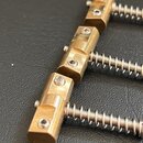 Zlatavý kovový most s třemi samostatnými bloky a připevněnými pružinami. Na každém bloku jsou vidět imbusové šrouby pro fixaci strun. Povrch vykazuje…