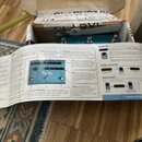 Otevřený kartonový obal, ve kterém je umístěno modré zařízení s několika ovládacími knoflíky a spínači. Před krabicí leží rozvinutý papírový návod s…
