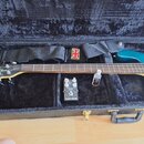 V černém plišovém kufru leží tyrkysová basová kytara, černý popruh s britskou vlajkou a efekt pedál. Na hlavě kytary je bílý nápis „Real Bass“ a menš…