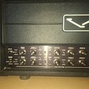Detailní pohled na ovládací panel kytarového zesilovače v černé barvě. Na panelu jsou nápisy INPUT, CHANNEL SELECT, GAIN I VOICING, GAIN II SATURATIO…