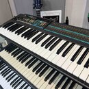 Dvě černé elektronické klávesové syntezátory umístěné nad sebou. Horní přístroj nese nápis MO-DX6 a má ovládací panel s modrými a červenými popisky.…