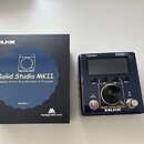 Na fotografii je kytarový pedál NUX Solid Studio MKII v tmavě modré barvě s černým ovládacím panelem a digitálním displejem. Vedle pedálu je umístěna…