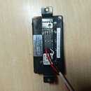 Černý plastový modul s elektronickými součástmi a připojenými červeným a bílým vodičem. Na modulu jsou viditelné nápisy EMG RA, GENUINE, (211017502),…