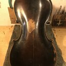 Zadní strana smyčcového nástroje, pravděpodobně violoncella, s tmavě hnědým lakem. Povrch vykazuje výrazné opotřebení, mnoho škrábanců a v centrální…