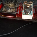 Dvě efektové pedály umístěné vedle sebe na černém povrchu. Levý pedál je černý s bílým grafickým vzorem a nápisem NEON CALL, má tři ovládací knoflíky…