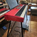 Červené digitální piano na černém kovovém stojanu s přiloženým sustain pedálem. Nástroj má klavírní klávesnici a ovládací panel s několika knoflíky a…