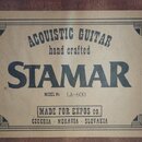 Fotografie detailu štítku z akustické kytary. Na béžovém podkladu je černý nápis ACOUSTIC GUITAR hand crafted STAMAR MODEL No LA-600. Dole je uveden…