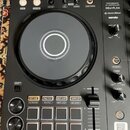 Černý DJ kontrolér s nápisy PERFORMANCE DJ CONTROLLER, DDJ-FLX4, rekordbox a serato. Zařízení obsahuje velký ovládací jog s bílým kruhem uprostřed, n…