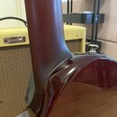 Detailní pohled na spoj hmatníku a těla tmavě červené elektrické kytary s lakovaným povrchem. V pozadí je vidět kousek zesilovače s nápisem Fender. N…