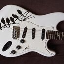 Bílá elektrická kytara s tvarem typu Stratocaster a bílým pickguardem. Na horní části korpusu je černý grafický motiv větve s pěti ptáky. Nástroj má…