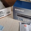 Fotografie zachycuje modrou krabici a bílý karton s produktem Shure BETA87A. Na bílém štítku je text „BETA87A Vocal Microphone Microphone pour la voi…