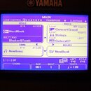 Detailní záběr na modrý LCD displej klávesového nástroje značky YAMAHA. Na obrazovce je zobrazeno menu MAIN s nastavením stylu HardRock, hlasů Concer…