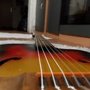 Detailní pohled na tělo a hmatník akustického nástroje, pravděpodobně basy nebo kytary. Povrch má červeno-žlutý přechodový odstín (sunburst) s černým…