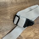 Šedý textilní popruh z tkaniny s černou plastovou přezkou a černým koženým zakončením. Na popruhu nejsou žádné viditelné značky ani nápisy. Povrch ma…