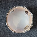 Fotografie znázorňuje snare drum z pohledu shora. Na průhledném bubenovém koži s nápisem Sonor a dopisky Made in Germany, 14" x 5.5" je umístěn kovov…