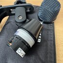 Na fotografii je černý dynamický mikrofon bez mřížky, vložený do černého držáku. Na spodní části kapsle je vidět bílý štítek s nápisy 8904, 094343 a…