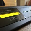 Detailní záběr na ovládací panel černého elektronického zařízení. Na povrchu je nápis YAMAHA, FM EXPANDER a TX7 TONE GENERATOR. Uprostřed je žlutý po…