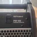 Detail horní části osobního monitoru značky Roland, model PM-100 PERSONAL MONITOR. Zařízení je šedé s černým ovládacím panelem, který obsahuje nápisy…