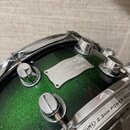 Detailní záběr na zelený buben s glitrovým povrchem a chromovaným obručem. Na korpusu je kovový štítek s nápisy MAPEX, Saturn Series a číslem 069704.…
