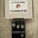 Černý efekt pedál s nápisy DevTech Audio a CUSTOM FUZZ. Na přední straně jsou tři stříbrné ovládací knoflíky s popisky VOLUME, TONE a SUSTAIN, v doln…