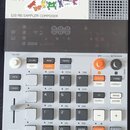 Šedý sampler s názvy K.O.II a nápisem サンプラー, pod kterým je text 128 MB SAMPLER COMPOSER. Zařízení obsahuje digitální displej, několik tlačítek v bílé…