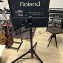 Černý kombozesilovač značky Roland umístěný na černém trojnožném stativu. Na horní hraně jsou ovládací prvky s nápisy POWER, PHONES, VOLUME, LOW, HIG…
