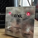 Kovový efekt pedál stříbrné barvy s broušeným povrchem. Na horní straně je černý nápis ENGL® a pod ním symbol s textem Tube Amp. Dole jsou dva stomeč…