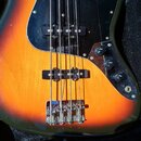 Detail elektrické basy v barvě sunburst s černým pickguardem, umístěná v pouzdře. Na těle jsou vidět dva snímače a tři ovládací knoflíky. Kovový most…