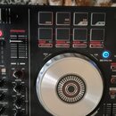 Detailní pohled na ovládací panel černého DJ kontroléru. Jsou vidět čtvercové podsvícené pady s nápisy SHIFT, SYNC, CUE a dalšími funkcemi jako SAMPL…