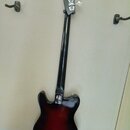 Zadní strana elektrické basy s černým krčkem a korýtkem v červeno-černém sunburst provedení. Na zadní straně korýtka je viditelný kovový kryt otvoru…